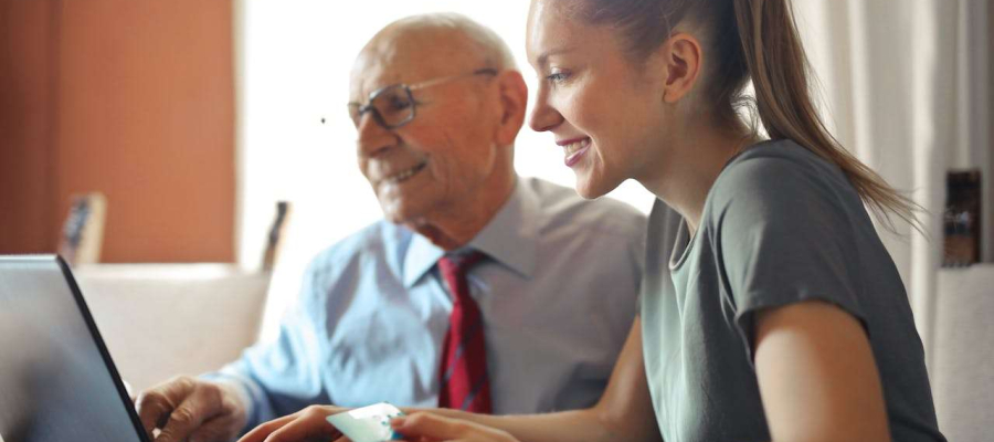 A woman is sitting with an older man, looking at a laptop. Both are smiling.